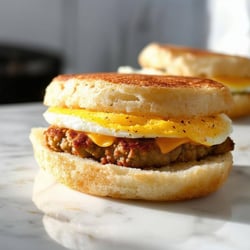 english muffin as the bread, with egg and sausage patty inside, like a mcdonalds item on a white marbel kitchen countertop